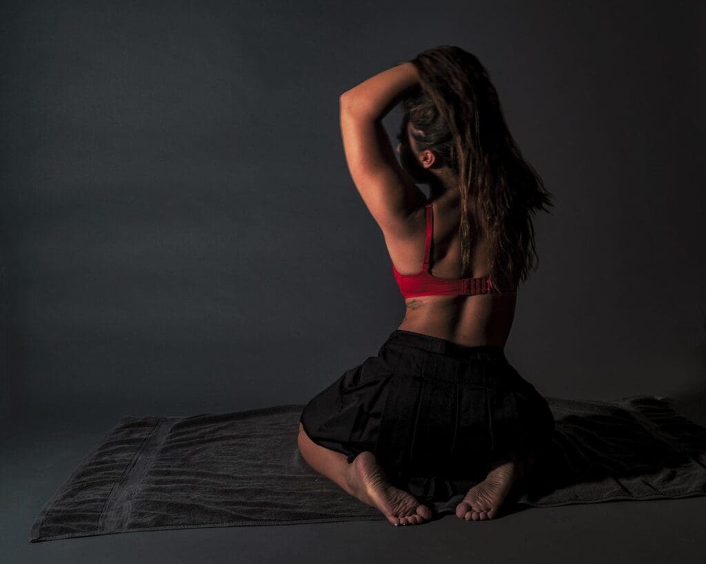 Danni kneels on a dark textile with one arm raised through her hair as angled light models her figure against the gray wall.